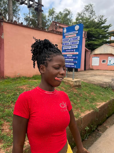 A young Black female with braided hair pulled into a bun, wearing a red shirt adorned with tiny rhinestones and a yellow skirt, is captured in a medium outdoor shot. She has her eyes closed and her tongue sticking out with what appears to be a lollipop or candy on it. Her expression suggests a playful or teasing demeanor.
In the background, a blue sign with white text indicates "DIOCESE OF NNEWI" and "BISHOP UZODIKE MEMORIAL SEMINARY (BUMSEM) NNEWI". Information on the sign includes "FOUNDED 1999" and details about classes and chapel worship times. To the right of the sign, a light pink building with a white framed window is visible, along with another building further back with a pitched roof. The scene is set against a backdrop of trees and a cloudy sky, with a concrete path and grassy area in the foreground, suggesting a location in an urban or semi-urban environment within Nnewi, Nigeria. An electrical pole with wires is also visible on the left side of the frame.