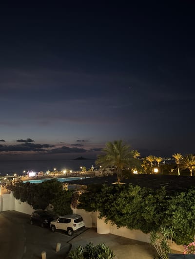 An elevated night-time view overlooks a coastal resort area in Capaci, Italy, with the Tyrrhenian Sea visible in the distance. The sky transitions from a deep blue at the top to lighter shades of purple and pink near the horizon, with scattered clouds. A small island or landmass with a structure on it is visible on the horizon. In the foreground, a swimming pool is illuminated, surrounded by lounge chairs and palm trees that are lit from below. Further back, a line of buildings with numerous lights suggests a resort or hotel complex. Two cars, a dark-colored compact car and a white SUV, are parked on a paved area in the lower left portion of the image, alongside a white wall and lush green bushes.