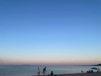 A serene beach scene unfolds at dusk in Nea Potidea, Greece. The sky transitions from a deep blue at the top to soft hues of pink and purple near the horizon, indicating either sunrise or sunset. The calm sea reflects the gentle colors of the sky. In the distance, a sailboat glides across the water, adding to the peaceful ambiance.

On the sandy shore, several individuals are enjoying the evening. Two women stand near the water's edge, one wrapped in a yellow towel, while another, also with a towel, appears to be talking to a third person. Further down the beach, a group is gathered under a blue umbrella, relaxing in beach chairs. A few people are wading in the shallow water, and the moon is visible in the sky, suggesting it is likely evening. The overall mood is one of tranquility and relaxation, with the soft lighting and gentle activity creating a calm atmosphere.