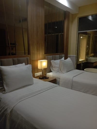 The image displays an unoccupied twin hotel room, characteristic of accommodations found in a city such as Batulawang, Indonesia. Two single beds, each equipped with a grey upholstered headboard, white duvet with subtle stripe patterning, and a single white pillow, are positioned parallel to each other. Between the beds, a light wood bedside table supports an illuminated rectangular table lamp with a cream-colored shade and a black remote control. Above the table, two white wall outlets are visible. The wall behind the beds features light wood paneling interspersed with vertical dark mirrored panels that reflect room elements. To the right, an opening or window offers a view into an adjacent bathroom, revealing a white freestanding circular bathtub in the foreground and a reflection of a white sink, faucet, and a wall-mounted hairdryer further back. The room is uniformly lit by warm artificial light. No individuals or active subjects are present.