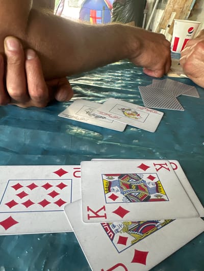 The image shows a close-up of a hand holding several playing cards on a blue, textured surface. The cards are laid out in a fanned arrangement, with two Kings of Diamonds prominently displayed. One King is upright, showing the full figure and the red diamond suit symbols. The other King is partially obscured, with only the top portion of its face and the red diamond symbols visible. A card with a "10" in red and diamond symbols is also visible, as is a card with a Joker.
In the background, a hand is visible reaching for a white cup with a red and blue logo. The logo appears to be that of Pepsi. Another hand is seen gripping the edges of some cards. The lighting suggests it is daytime. The scene appears to be an informal setting, possibly a game of cards being played at a table. The blue surface could be a tablecloth or a plastic covering. There are no strong indicators of the weather or specific time of day beyond daylight.