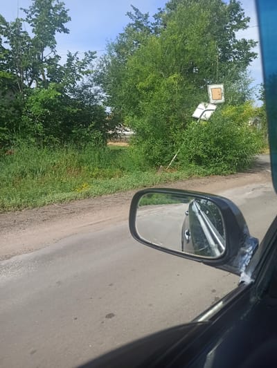 The image shows a roadside scene with lush green vegetation. In the background, two road signs are visible amidst the dense foliage. One sign is square with an orange-yellow interior, and the other is a white rectangular sign with a black arrow indicating a curve to the left. The road appears to be paved with asphalt and has some dirt and gravel on its shoulder. A side mirror of a vehicle is prominently featured in the foreground, reflecting the road ahead and the side of the car. The sky is blue with some clouds. The overall impression is of a rural or semi-rural setting, possibly in Novoselivka, Ukraine, given the context.
