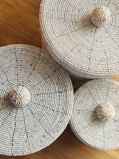 Three round, lidded containers, covered in small, off-white beads, are arranged on a wooden surface. Each lid has a small, spherical knob made of the same beads at its center. The containers are placed close together, with the lid of the largest container visible in the upper portion of the image. The other two containers are positioned below and to the left and right of the largest one, with their lids facing upwards. The wooden surface has a visible grain pattern. The scene is an indoor setting and does not provide any information about the city of Maitland, United States of America.