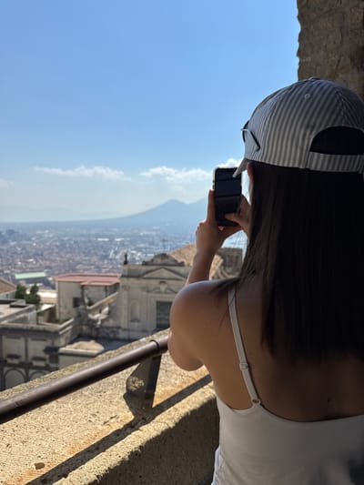 In this vertical medium shot, the back of a young woman wearing a striped baseball cap and a white tank top is captured as she takes a photo with her phone. She is standing at a viewpoint, looking out over a sprawling city with a mountain in the distance. The city appears to be Naples, Italy, with its dense urban landscape and historical architecture.
The woman's dark hair falls over her shoulders, partially obscured by her cap. Her hands are visible, holding her smartphone horizontally as she frames a shot of the cityscape. The sunlight casts shadows on the stone ledge she is leaning against, suggesting it is daytime, likely late morning or early afternoon. The sky is a clear, bright blue with a few wispy clouds.
The background reveals a panoramic view of the city, with many buildings and the distinct silhouette of a mountain range. The architecture suggests a Mediterranean setting. The overall mood is one of sightseeing and enjoying a scenic vista. There is no visible text in the image.