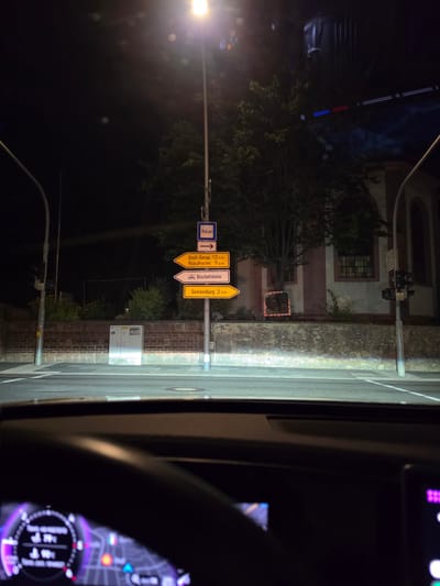 The image shows a street scene at night in Bischofsheim, Germany. A lamppost with street signs is prominently featured. The signs indicate directions to "Polizei" (Police), "Groß-Gerau 13 km", "Nauheim 9 km", "Bischofsheim", and "Gustavsburg 2 km". The scene is viewed from inside a vehicle, with a dashboard and a digital display visible in the foreground. A brick wall and a building with windows are in the background. The lighting suggests it is nighttime, with streetlights illuminating the area. There are no people visible in the image.
