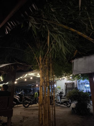 The image depicts an outdoor scene, likely a courtyard or open-air dining area, at night. Several motorcycles and scooters are parked on a dirt or gravel surface. Strings of lights are strung above, providing illumination to a covered structure with a tin roof. A person with a backpack is visible near a parked motorcycle. A bamboo structure stands prominently in the foreground. The environment appears to be an establishment serving food or drinks, indicated by the presence of tables and chairs under the covered area. The location in Perugia, Italy, cannot be confirmed from the image.