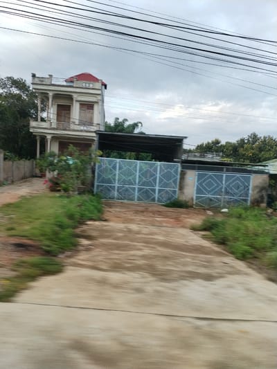 The image depicts a residential scene in Đăk Xú, Vietnam. A two-story house with white exterior, red roof, and colonial-style architectural details, is visible on the left. A gated entrance sits to the right. The gate appears to be made of a light blue metal with a geometric pattern. A covered structure is located behind the gate, with various trees and vegetation present in the background. Overhead, numerous power lines stretch across the sky, which appears overcast. The foreground is comprised of a weathered, concrete driveway and patches of green vegetation. The image appears to have been taken while in motion, resulting in some blur.
