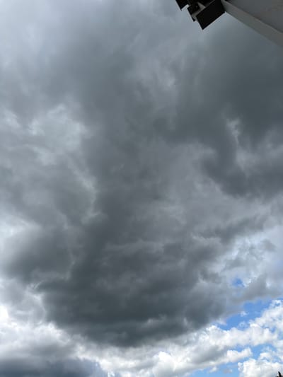 The image shows a dramatic sky filled with dark, ominous storm clouds. The clouds are dense and appear to be moving, with varying shades of gray and white. In the upper right corner of the image, a portion of a modern building's structure is visible, casting a shadow on the sky. The building appears to be made of metal and concrete. The lower part of the image reveals patches of clear blue sky with some lighter, fluffier clouds, indicating that the storm might be localized or passing. The scene suggests a turbulent weather event over the city of Janowice, Poland.