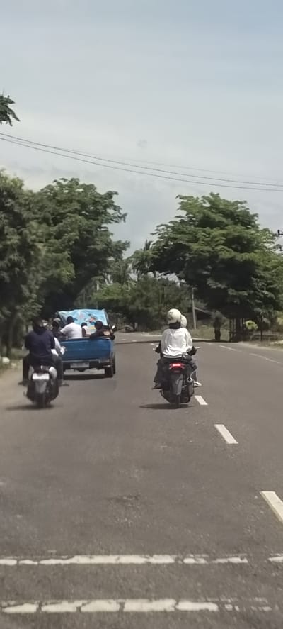 The image depicts a road scene in Aluekeujruen, Indonesia. Two motorcycles and a small truck are traveling in the same direction on a paved road with lane markings. The truck appears to be carrying multiple passengers in its bed. Trees line the roadside. The sky is overcast.