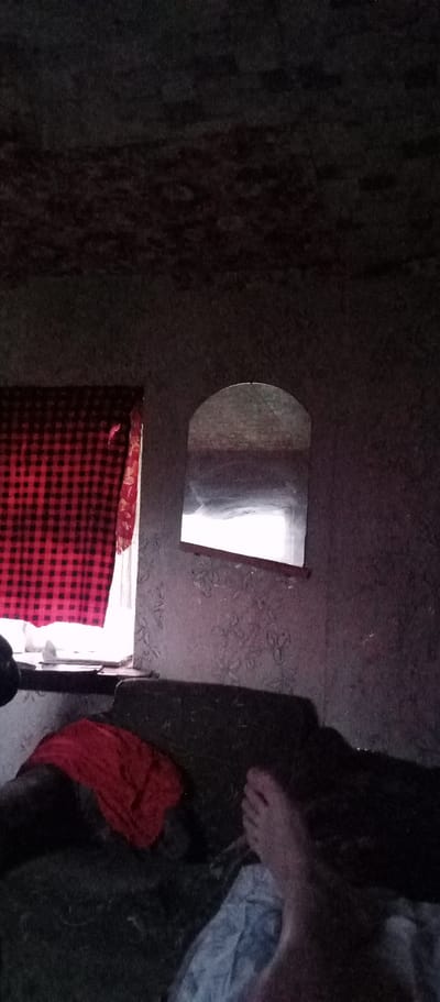 An interior view of a dimly lit room with a bed, a window covered by a red and black plaid curtain, and a mirror on the wall. The ceiling appears to be textured, possibly with mold or discoloration. A person's leg and foot are visible on the right side of the frame, resting on what appears to be bedding or a mattress. A red cloth is draped over a dark-colored surface, likely part of the bed. The overall atmosphere is somber and unkempt, suggesting a lack of recent maintenance or a very basic living condition. The location is identified as Marinovka, Kazakhstan, but no specific geographical features or contextual clues within the image support this claim beyond the provided metadata.