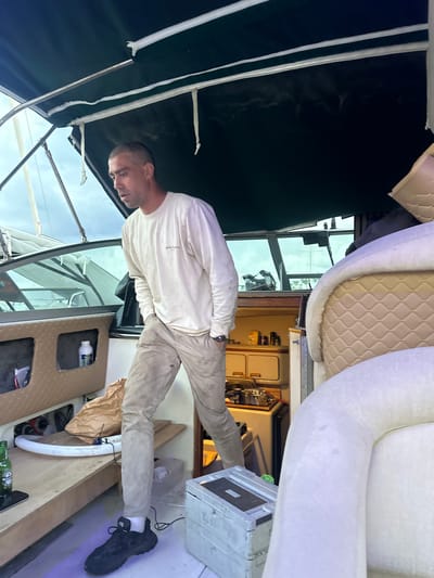 A man stands on the deck of a boat. The boat is docked in Vallauris, France. He is wearing a long-sleeved white shirt and khaki pants. The boat's interior includes white padded seating, cabinetry, windows, and a dark awning. Other details visible include tools, bottles, and a wooden deck.