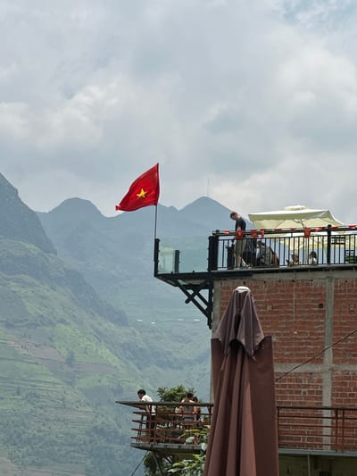 The image shows a scene from Mèo Vạc Commune, Vietnam. A Vietnamese flag is prominently displayed against a backdrop of mountainous terrain and a cloudy sky. A structure with a brick building and a glass-floored balcony extends from the mountainside. People are on the balcony, and a brown umbrella partially obscures the view.