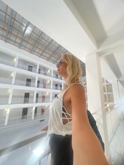A woman with blonde hair and sunglasses, wearing a white sleeveless top and black pants, stands on a balcony overlooking a multi-story building with open corridors. The building has white railings and dark doors on each level. The woman is holding a black bag and extending her right arm, possibly taking a selfie. The environment appears to be an interior courtyard or atrium of a hotel or residential complex. The lighting is bright, with natural light entering from above and artificial lights illuminating the building's facade. The location is identified as Giachrypsh, Abkhazia.