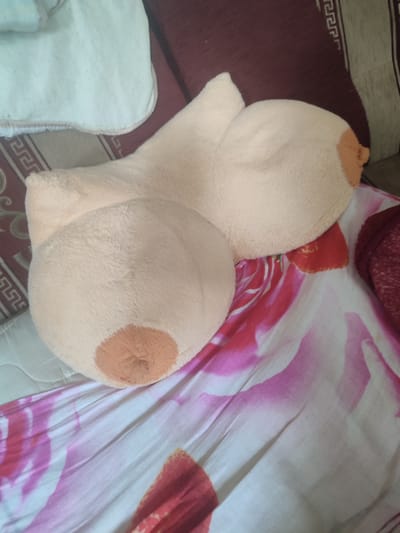 A plush toy shaped like breasts, with a light peach color and darker peach nipples, is placed on a bed. The bed is covered with a white and pink floral patterned sheet. In the background, there is a burgundy upholstered headboard with a decorative pattern, and a light-colored blanket or pillow. The scene appears to be indoors, likely a bedroom. There is no indication of the location being in Velikii Krai, Russia.