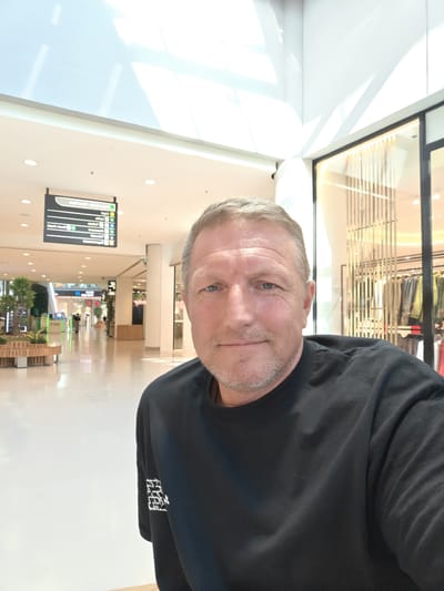 A middle-aged Caucasian man with short, blonde hair and a light beard smiles into the camera in a selfie. He is wearing a black t-shirt. The man is inside a modern, brightly lit shopping mall. In the background, there is a long, wide corridor with a polished floor. Signs are visible hanging from the ceiling, indicating directions and store information. To the right of the man, there is a storefront with clothing displayed. To the left, there are planters with greenery and a bench. The architecture of the mall features large windows and high ceilings with a modern lighting system. The scene appears to be in the city of Nola, Italy, as indicated by the signage.