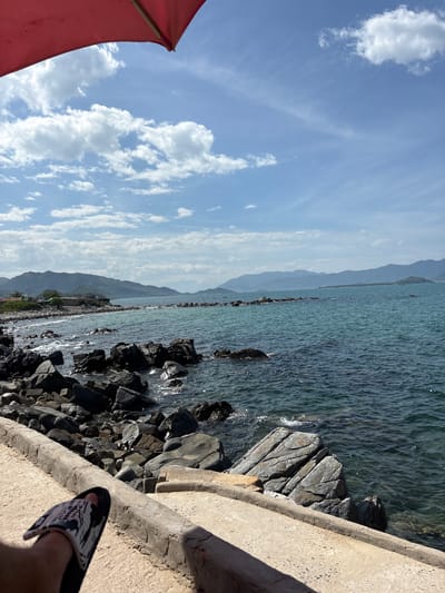 A person's foot wearing a sandal rests on a concrete ledge overlooking a rocky coastline in Vĩnh Lương, Vietnam. The sea is calm and blue, with gentle waves lapping against the dark rocks. In the distance, lush green mountains form a scenic backdrop against a sky filled with white, fluffy clouds. A portion of a red umbrella is visible in the upper left corner of the frame. The overall impression is one of a peaceful day at the beach.