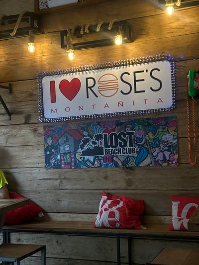 The image shows a scene inside a restaurant in Montanita, Ecuador, located at coordinates -1.826 latitude and -80.752 longitude. The restaurant is decorated with a rustic wooden wall, a long wooden bench, and a few red and white heart-shaped pillows on it. Two bright lights are attached to the ceiling with rustic-looking ropes. A red phone is plugged into the wall, and a red and orange cord dangles from the phone. There is a white sign with the words "I LOVE ROSE'S MONTANITA" in black lettering. Below the sign is a colorful poster of a "LOST BEACH CLUB" and a partially visible man wearing a yellow shirt seated at a table with a partially visible wooden chair. There is a small square wooden table in front of the bench. The room is dimly lit with the lights casting a warm glow.