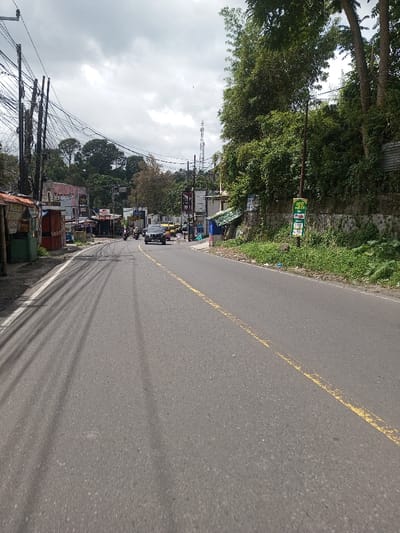 The image presents a street view in Gubbisamamidi, India, under an overcast sky. The foreground features a two-lane asphalt road marked with a continuous yellow line down the center and a continuous white line along the left edge. Long, parallel shadows from overhead utility lines are cast across the left portion of the asphalt.
On the left side of the road, multiple utility poles are visible, heavily strung with a dense network of black electrical and communication wires. Below these poles, a series of simple, one-story commercial structures, some appearing as open-front kiosks or stalls, line the roadside. Multiple indistinct human figures are present near these establishments.
On the right side of the road, an elevated, heavily vegetated embankment rises, supported by a stone and concrete retaining wall. A green pole with multiple stacked green rectangular signs, one clearly labeled "100 METER," is positioned along this side.
Vehicular traffic is present in the mid-ground, including a dark-colored passenger car traveling away from the viewer in the left lane. A motorcycle is visible behind it, and further down the road, additional vehicles, some with yellow coloration, are present. The background consists of a dense treeline on undulating terrain and a tall, lattice-structured communication tower extending into the cloudy sky.