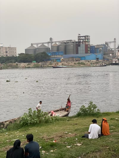 The image depicts a bustling river scene in Tarabo, Bangladesh, under an overcast sky. The wide river forms the central element, its surface reflecting the subdued light and featuring scattered patches of green floating vegetation, likely water hyacinths.
On the far bank of the river, an extensive industrial complex dominates the background. This complex includes numerous towering grey silos, large multi-story buildings with blue and grey facades, and a network of intricate metal structures, possibly cranes, gantries, or conveyor systems, indicative of a processing or manufacturing plant. A long vessel, potentially a cargo boat, is visible on the river near this industrial bank, suggesting riverine transport. Further in the distance, a multi-story building, possibly residential or commercial, can be seen.
The near bank of the river features a green, grassy slope descending to the water's edge. This area appears somewhat untended, with varied grass textures and scattered debris or litter. Several individuals are present on this bank. In the immediate foreground, two people are observed crouching or sitting low; one wears a white t-shirt and dark trousers, and another is partially covered by an orange garment. Further back on the bank, another person in a light-colored shirt stands near some low-lying foliage.
A small, traditional wooden boat is positioned close to the grassy bank in the mid-ground. A person, possibly wearing striped clothing, is visible within this boat, either standing or engaged in an activity. The soft, even lighting suggests it is daytime, but an overcast or hazy sky rather than direct sunlight. No distinct text is clearly legible on any signs or buildings within the image.