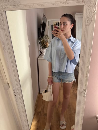 A woman is taking a mirror selfie in a room. She is wearing a light blue short-sleeved button-up shirt, denim shorts, and slide sandals. She is holding a cream-colored handbag. A mirror with a distressed white frame is in the foreground. The room appears to be a bedroom or dressing area, with a wooden floor, a closet, and a television mounted on the wall. The lighting is bright and natural. The specific location in Targovishte, Bulgaria is not determinable from the image.