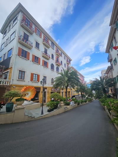 The image captures a street scene in Phú Quốc, Vietnam, likely within a resort or entertainment complex designed with European-inspired architecture. The foreground shows an asphalt road curving to the right, bordered by raised planters filled with lush green foliage and flowering plants.

On the left, a multi-story building with a light-colored facade features numerous balconies adorned with red shutters and decorative figurines. An oversized, abstract orange sculpture resembling a stylized flower is prominently displayed in front of this building, situated on a raised platform with steps. A black lamppost stands nearby.

The street is lined with more buildings in a similar architectural style, painted in various pastel colors like pink and yellow, with palm trees interspersed throughout. The buildings have balconies, arched windows, and decorative detailing. The sky above is a vibrant blue with streaks of white clouds. The overall impression is one of a well-maintained, picturesque street designed to evoke a Mediterranean or European ambiance. There are no people visible in the image.