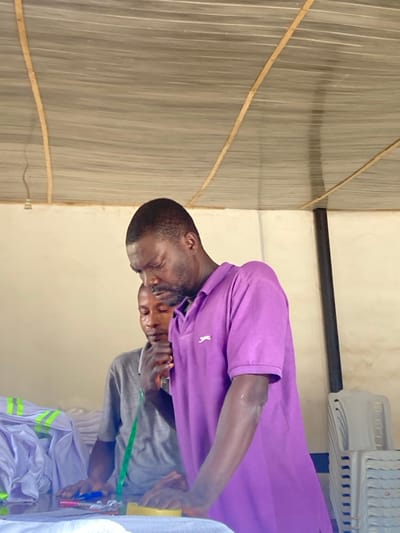 In an indoor facility in Agege, Nigeria, two adult males are observed. The prominent figure in the foreground, a man with dark hair and a short beard, wears a purple polo shirt featuring a white feline logo on the left chest. He is leaning forward, directing his gaze and hands toward a white fabric spread on a surface. Adjacent to his hands are a yellow sponge-like object, a red-capped pen, and a blue tool. Behind and to the left, a second man in a grey t-shirt, with a green measuring tape draped around his neck, also looks down intently at the work surface; his right hand rests near his chin. To the left of the men, numerous stacks of neatly folded white garments, some exhibiting yellow-green reflective bands, are visible. On the right side of the frame, a stack of light-colored plastic chairs is present. The environment consists of light-colored walls and a ceiling composed of corrugated material supported by wooden beams. A black vertical support or pipe is situated on the right, and an exposed light fixture hangs from the ceiling on the left.