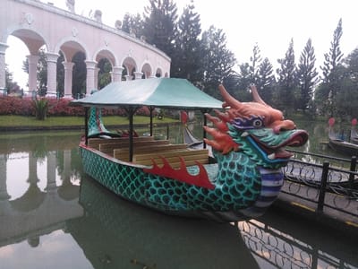 This image depicts a brightly colored, unoccupied dragon-headed pedal boat floating in a calm body of water within a recreational park in Ciudad de San José, Costa Rica. The boat, positioned prominently in the foreground, features a vivid green scaled hull, red fin-like adornments, and an elaborate multi-colored dragon head at its bow, rendered in orange, blue, green, and purple hues. A green canopy covers the passenger area, which contains light-colored wooden bench seating.
To the left, a light-pink, neo-classical arched structure with multiple white columns spans the background, reflecting partially in the water. Below this structure, a landscaped bank exhibits dense red and green foliage. In the mid-ground on the right, several other smaller, unoccupied boats, resembling stylized gondolas, are partially visible on the water, moored near a dark metal railing that borders the water's edge. A dense line of tall, dark green coniferous trees forms the distant background under an overcast sky, suggesting an outdoor park setting. No individuals are present in the scene.