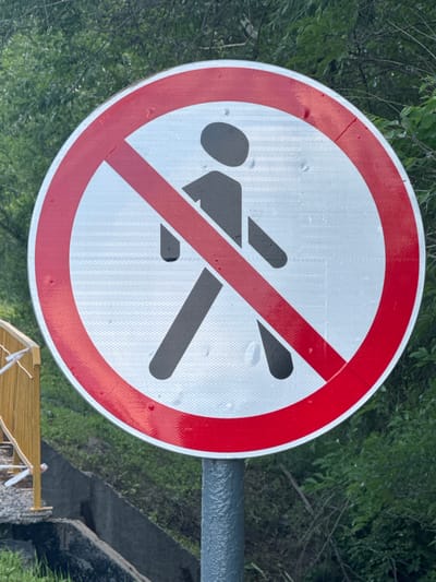 The image shows a close-up of a circular traffic sign with a red border and a red diagonal line crossing a silhouette of a person. The sign is mounted on a gray pole. The background consists of lush green foliage and trees. To the left of the sign, a wooden fence with yellow posts is partially visible. The ground appears to be a mix of gravel and concrete, with some grass. The sign is positioned outdoors in what appears to be a natural or semi-natural environment, possibly a park or a roadside. The overall scene suggests a "no pedestrians" or "pedestrian crossing prohibited" sign. The image does not contain any recognizable landmarks that would definitively place it in Novosemeykino, Russia.
