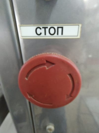 The image shows a close-up of a red emergency stop button on a metallic surface. Above the button, a rectangular label with a white background and black text reads "СТОП" which is the Russian word for "STOP". The button itself is circular with a textured edge and features three indented arrow shapes pointing in a clockwise direction around its circumference. The metallic surface appears to be part of a machine or industrial equipment, with a brushed texture. The lighting is overhead and casts a slight shadow. There are no people or animals visible in the frame. The image does not provide any information about the city of Tervel, Bulgaria.