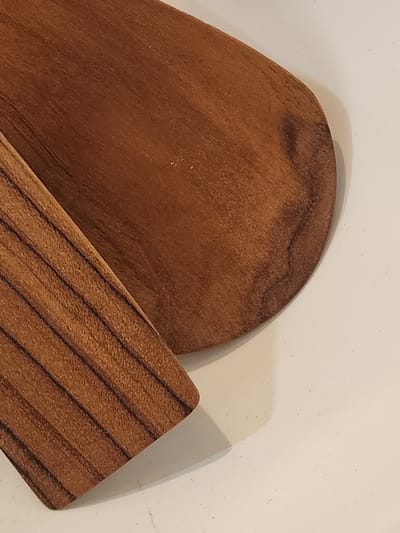This is a close-up, overhead shot of two wooden objects with a light-colored, textured background. The objects are a warm, reddish-brown color and appear to be made of polished wood, with visible grain patterns.
The larger object, positioned on the left, has a curved edge on its top right. Its surface displays swirling grain patterns and some darker, irregular markings. It lies flat against the background.
The second object, situated in the lower right quadrant, is thinner and has a distinct linear grain pattern, appearing to be composed of alternating lighter and darker stripes running diagonally. This object is angled on top of the larger wooden piece, creating a layered effect.
The lighting suggests a diffused light source, casting a soft shadow from the objects onto the background. There are no people or text visible in the image. The setting appears to be indoors, possibly a tabletop or a plain surface, given the neutral background and controlled lighting. The overall impression is one of simple, natural materials presented in a still-life arrangement.