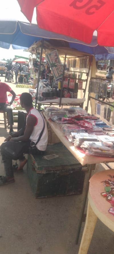 The image depicts an outdoor market scene in Isukha North ward, Kenya, under bright daylight. In the immediate foreground, a male with dark skin is seated on a dark green metal trunk. He wears a white sleeveless shirt with red trim, dark trousers, and a black shoulder bag resting on his lap. To his right, partially visible, is a small, light-colored round table displaying multiple colorful keychains or small locks.
Behind this individual, a street vendor stall is set up, shaded by two large parasols: one red (partially visible at the top right) and one blue and white (at the top left). The stall features a metal framework from which various packaged items, primarily phone covers and accessories, are suspended. A prominent vertical white sign with "M-PESA" printed in green is attached to the stall's structure. Below the hanging merchandise, a table draped in white fabric displays stacks of various packaged goods, including stationery and small electronics accessories.
To the left of the central seated male, a second male is visible, seated on a stool, wearing a red t-shirt and dark trousers. Behind him, on the ground, a pile of large green produce, possibly watermelons, is arranged for sale. The background shows a bustling street scene with several blurred figures, some standing near motorcycles, and a yellow matatu (public transport van) partially visible further in the distance. The ground surface in the foreground appears to be paved, transitioning to unpaved or dirt in the background.