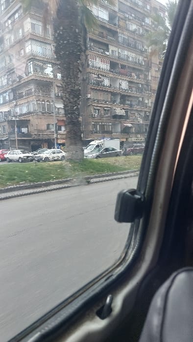 The image depicts an urban street scene in Al-Qaboun Municipality, Syria, captured from the interior of a moving vehicle. The right side of the frame is occupied by the vehicle's light-colored window frame, a dark window latch mechanism, and a portion of a gray seat. The road surface in the foreground shows motion blur. Beyond the road, a grassy median strip with a concrete curb is visible. Several vehicles are stationary or parked on a dirt shoulder adjacent to the median. These include at least two white older-model sedans, one with visible markings on its side, a silver sedan, a white box-style van with blue details, and a red car further in the distance. A large palm tree with a thick, textured trunk stands prominently in the midground. In the background, a multi-story, concrete building, likely residential or mixed-use, occupies a significant portion of the frame. This building features multiple windows, enclosed balconies, and numerous external air conditioning units across its facades, indicating an aged urban structure. The sky is overcast. No individuals are clearly identifiable.