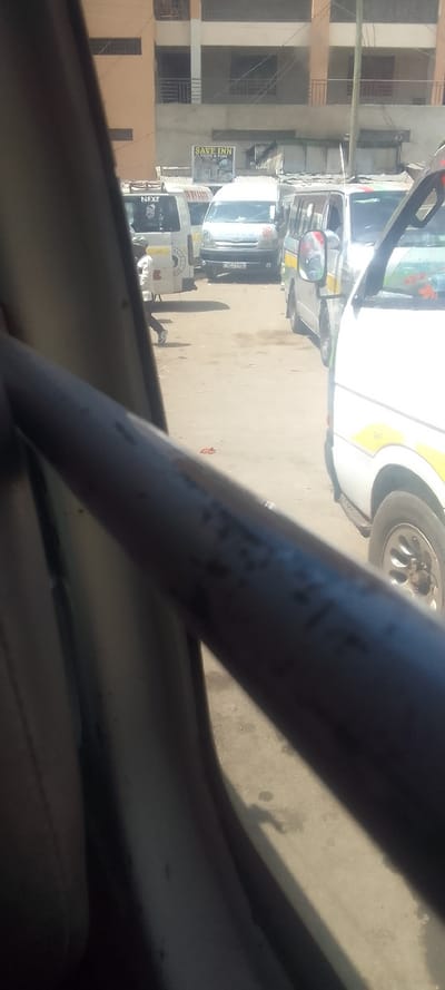 The image presents an urban street scene in Naivasha, Kenya, captured from the interior of a vehicle. A dark, tubular metal bar diagonally traverses the lower center, partially obscuring the view, with a light-colored vehicle interior panel visible to its left. The ground is unpaved, appearing as light brown or grey dirt.

In the middle ground, several white minibuses, commonly identified as matatus, are positioned. One matatu on the far left displays "NEXT" on its side, along with a circular emblem. A male individual, dressed in a light shirt and dark trousers, walks from left to right behind this matatu. A central matatu prominently displays the license plate "KC J 278S". To the right, another matatu, white with yellow stripes, is partially visible.

The background features multi-story buildings constructed from a combination of light brown and grey materials, with visible balconies and window openings. Below these, a row of single-story structures with corrugated metal shutters is present. A prominent commercial sign for "SAVE INN CHIPS & FISH" is mounted on one of these lower structures. Utility poles and overhead electrical or communication lines are visible against the buildings. The scene depicts a commercial or transport hub during daylight hours, with vehicles appearing stationary or moving slowly.