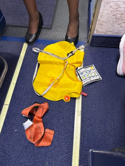 The image shows a close-up view of a section of an airplane cabin floor. On the dark blue carpet, there is a bright yellow life vest, a red seatbelt, and a folded safety instruction card. The life vest is partially open and has straps. The seatbelt is unbuckled and lying in a loop. The safety instruction card shows diagrams of how to use safety equipment. Two pairs of legs are visible: one pair wearing black high-heeled shoes and dark stockings, and another pair wearing pink sneakers. The presence of safety equipment and the cabin floor suggest an airplane. The question asks to locate this scene in Palma de Mallorca, Spain, but there is no visual information in the image that can confirm this specific location. Therefore, a precise location cannot be determined from the image alone.