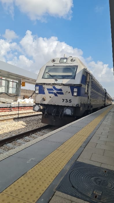 A daylight vertical photograph depicts a stationary Israel Railways locomotive, identified by the number "735" and the company's silver-grey and blue livery, at a train station platform in Rishon LeZion, Israel. The front of the locomotive, featuring its operational lights and insignia, is positioned on the track directly adjacent to the concrete platform. The platform includes a yellow tactile warning strip with raised dots along its edge and a dark, circular manhole cover visible in the foreground. To the left of the train, multiple railway tracks with gravel ballast are present. A modern white canopy structure extends over a portion of the platform in the upper left. Further in the background, an area with construction materials and red safety barriers is visible next to the tracks, with multi-story residential buildings discernible in the distance. The sky above is blue with scattered white clouds. No individuals are visible within the frame.