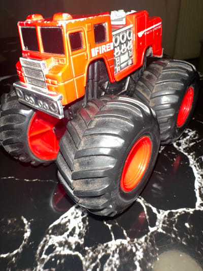 The image shows a close-up, eye-level, indoor shot of a red and orange toy monster truck fire engine. The truck is highly detailed, featuring a chrome grille, large black tires with red rims, and the word "FIRE" emblazoned on the side. There are also markings that read "SEARCH & RESCUE" on the rear fender. The truck is positioned on a glossy, black surface with a white marbled pattern. The lighting is somewhat dim, casting shadows that highlight the texture of the tires. The background is dark and out of focus. There is no indication of the city of Fulda, Germany, in the image.