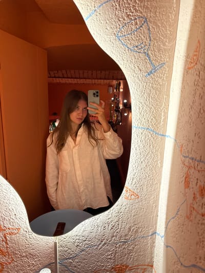 A young adult female is captured in a mirror selfie within an establishment located in Vracar Urban Municipality, Serbia. The woman, with long brown hair, wears a light-colored, loose-fitting collared shirt over dark clothing. She holds a light-green smartphone with three camera lenses in her right hand, positioned to capture her reflection; a silver ring is visible on her right ring finger. Her gaze is directed towards the camera.
The mirror itself is irregularly shaped, with an illuminated border, and reflects an interior space painted a uniform warm orange-red hue. Visible in the reflection are a plain orange wall to the left and, through an arched opening on the right, a dimly lit bar area with various bottles on shelves and warm-toned light fixtures. The ceiling in the reflected area appears to have exposed wooden beams or similar architectural detail.
The wall surrounding the mirror is textured with a rough, stucco-like finish and is adorned with various hand-drawn-style illustrations in blue and orange. These include a blue outline of a wine glass and abstract wavy lines, alongside orange outlines of fruit slices and other undefined shapes. A small, circular light fixture on a slender stand is visible at the bottom edge of the mirror, with a dark, rectangular object (possibly a remote control) resting on the surface directly below the mirror's reflective surface.
