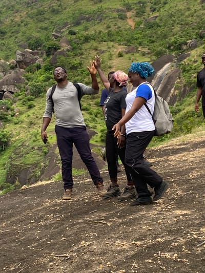 A group of four individuals stand on a rocky terrain, seemingly on a mountain or high elevation, with a lush green landscape visible in the background.
The foreground shows the textured rocky surface they are standing on. The landscape in the background includes vegetation and rock formations.
From the original caption: The location of this image is likely in the vicinity of Ganawuri, Nigeria.