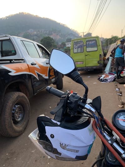 The image captures a vibrant street scene in Freetown, Sierra Leone, viewed from a slightly rotated perspective, likely from atop a motorcycle. In the immediate foreground, a white motorcycle helmet rests on the handlebar of a bike, partially obscuring a speedometer. The bike's rearview mirror reflects a clear, bright sky. The helmet bears a faded, distinctive sticker.

Beyond the motorcycle, a bustling street unfolds. A light green van is prominent in the mid-ground, with the word "TIME" visibly painted or stickered on its rear window. Further down the street, a black and white pickup truck is parked, adorned with a large orange and white "4G" graphic and other text that appears to read "3000...". Two spare tires lean against the truck on the ground, one faintly showing "GOODYEAR".

Several people are congregated near the green van and the pickup truck, some appearing to be engaged in market activities with items, possibly clothes, laid out on the ground. The background reveals a hilly urban landscape with numerous buildings nestled into the terrain. The warm, soft lighting suggests late afternoon or early evening, with the sun casting a golden glow on the horizon. The overall scene depicts a lively urban environment with commercial activity.
