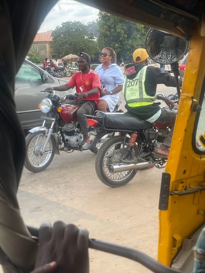 In Galadimawa Village, Nigeria, this image captures a street scene with multiple motorcycles and people. The foreground shows a blurred hand gripping a dark steering mechanism, likely from a yellow auto-rickshaw, with a portion of its yellow body visible on the right. In the middle ground, two motorcycles are prominent. The motorcycle on the left is red and driven by a man wearing a red t-shirt with a white logo and a black baseball cap. He is looking to his right. Behind him, on the same motorcycle, a woman wearing a light blue collared shirt and sunglasses is seated. To the right of this motorcycle, another motorcycle is visible, with a driver wearing a yellow baseball cap, a black hooded jacket over a lime green reflective vest labeled "207 FCT ARSRA," and dark pants. The background reveals trees, buildings with terracotta roofs, and a cloudy sky. A small stall with goods displayed on shelves is situated behind the motorcycles. The road surface appears to be asphalt or packed dirt.
