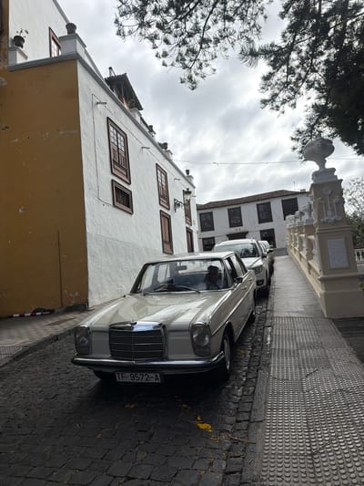 This is a street-level, eye-level, medium shot photograph taken outdoors on a cloudy day in Icod De Los Vinos, Spain. The foreground features the front half of a vintage, light-colored sedan parked on a cobblestone street. The car has a chrome grille, a hood ornament, and a license plate that reads "TF-9572-A". A person's head and shoulders are visible through the driver's side window. To the right of the car, a raised, textured walkway with tactile paving runs alongside a decorative, ornate white balustrade and a light-colored building. Behind the vintage car, a white SUV and another dark-colored vehicle are parked, partially visible. The background shows a multi-story building with white walls and dark-framed windows, characteristic of Canary Island architecture. A large tree with dark green foliage partially obscures the sky at the top of the image. The left side of the image is dominated by a building with a section of yellow-painted wall adjacent to a white wall with dark windows. The street appears to be narrow, with a sidewalk on the right.