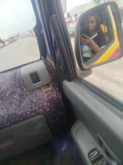 In Uselu, Nigeria, a person is seen taking a selfie in the side mirror of a vehicle. The person is wearing a black t-shirt with a colorful graphic design and has braided hair. The interior of the vehicle shows a furry texture on the door panel and a grey air vent. Outside the vehicle, a road with other vehicles and buildings can be seen, suggesting a bustling urban environment. The weather appears overcast.