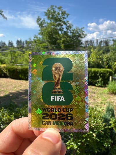 A hand is holding a rectangular sticker with a holographic background. The sticker features the FIFA World Cup 2026 logo, which includes the number "26" in large green digits and the FIFA logo. Below that, it reads "WORLD CUP" and "CAN MEX USA". The background of the sticker is a repeating pattern of the word "PANINI" in various colors and styles, interspersed with small FIFA logos. The sticker is shiny and catches the light, creating a shimmering effect. The background behind the sticker is blurred, showing greenery, trees, and a blue sky with clouds, suggesting an outdoor setting. The sunlight suggests it is daytime. The scene is likely in Prisovo, Bulgaria, given the location context.