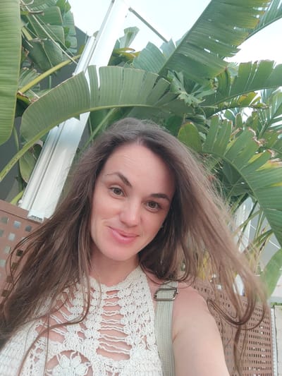 A medium shot captures a woman from the chest up, posed in front of a backdrop of large, green leaves, likely belonging to a banana tree or similar tropical plant, common in the Mediterranean climate of El Campello, Spain. The woman has long, brown hair that is slightly windswept, framing a face with brown eyes and a faint smile. She is wearing a cream-colored, crocheted top with a distinctive openwork pattern, and a light-colored strap is visible over her right shoulder. Behind her, a white, cylindrical structure, possibly part of a building or light fixture, stands vertically. The foreground includes a brown, lattice-like fence or seating area. The lighting suggests an outdoor setting, likely during daytime.