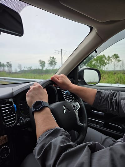 A man is driving a car. He has both hands on the steering wheel and is wearing a smartwatch. The car appears to be on a highway, and the background shows trees. The location is Аур, Russia, as the picture location implies a location in that city.