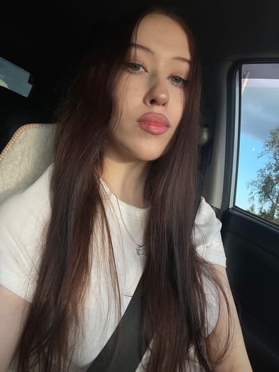 A young woman with long, dark brown hair is captured in a selfie from a slightly elevated angle, likely taken inside a vehicle. She is wearing a white t-shirt and a subtle necklace with a pendant. Her makeup is natural, highlighting her eyes and lips. The background shows the interior of a car, with a glimpse of the sky and trees through the sunroof, suggesting it's daytime. The lighting indicates it might be late afternoon or early evening. The overall mood is relaxed and casual. The image appears to be from Gavrilovka, Russia.