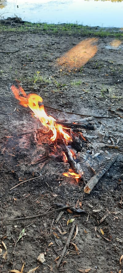The image shows a campfire on a muddy ground, likely near a body of water, given the reflection of light on the left side. The fire is burning with orange and yellow flames, illuminating the surrounding ash and burnt wood. The ground is covered with dirt, leaves, and small twigs, suggesting an outdoor setting, possibly a forest or a park. The lighting suggests it might be daytime, but the shadows are not very pronounced, and the overall tone is somewhat muted, hinting at an overcast sky or late afternoon. There are no people visible in the image, and the focus is solely on the campfire and its immediate surroundings. The scene evokes a sense of wilderness and a moment of quiet solitude. There is no visible text in the image.
