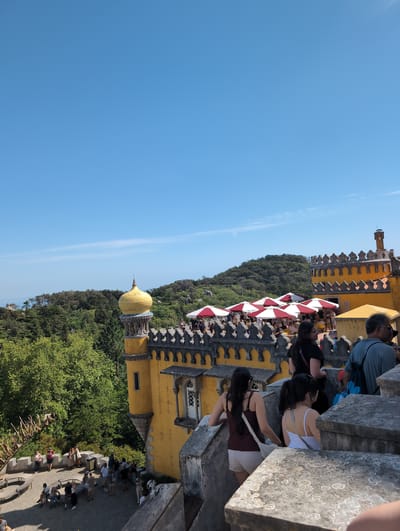 The image shows an outdoor scene in Sintra, Portugal. The prominent feature is a section of the Pena Palace, with its yellow walls and decorative architectural details. The sky is blue. People are visible on the palace's terrace and in the surrounding areas, suggesting they are tourists or visitors. The landscape around the palace is characterized by lush green vegetation, including trees. Red and white awnings are visible near the crowd.