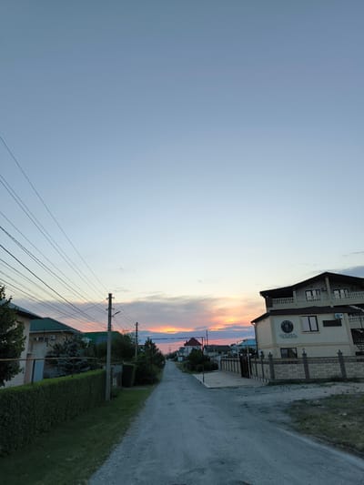 The image depicts a street scene in a residential area, likely during sunset given the warm hues in the sky. A paved road runs through the center of the frame, flanked by houses and greenery. On the left, a neatly trimmed hedge borders a house with a green roof and visible electrical wires overhead. On the right, a multi-story building with a balcony and what appears to be a business sign with Cyrillic text is prominent. The sign reads "ЧЕХОВЪ" and "ОТДЫХ" which translates to "Chekhov" and "Rest/Leisure". The sky above is a gradient of pale blue fading into the orange and pink tones of the sunset, with some scattered clouds. The overall mood is tranquil and serene. The presence of power lines and the architecture suggest a moderately developed, possibly suburban or rural, setting. The time of day is clearly evening, and the weather appears clear. No people are visible in the frame.
