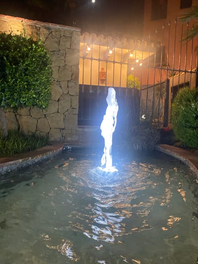The image depicts an outdoor fountain at night, with the water illuminated by a bright light source, creating a luminous effect in the jet. The fountain is situated within a courtyard-like setting, bordered by a stone wall on the left and a body of water on the right. Behind the stone wall, a building facade is visible, featuring large windows with decorative metal grilles. String lights hang across the facade, casting a warm glow. In the foreground, a neatly trimmed green bush is visible. The overall scene suggests a tranquil and inviting atmosphere, possibly a patio or garden area. The time of day is clearly night, and the lighting suggests no particular weather conditions. No visible text is present in the image.